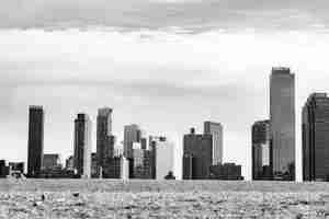Black-and-white city skyline along a waterfront, with tall buildings rising above a calm, pebbled shore under a cloudy sky
